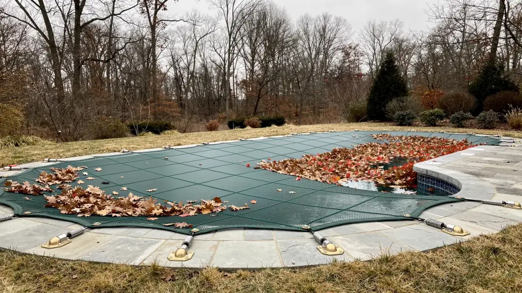 Inground pool closed for winter under a green mesh safety cover with fallen leaves